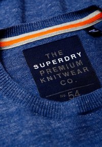Blauer Strickpullover mit Rundhalsausschnitt und ribbeligem Kragen. Etikett mit dem Markennamen "Superdry" und einem Fischgrätenmuster.