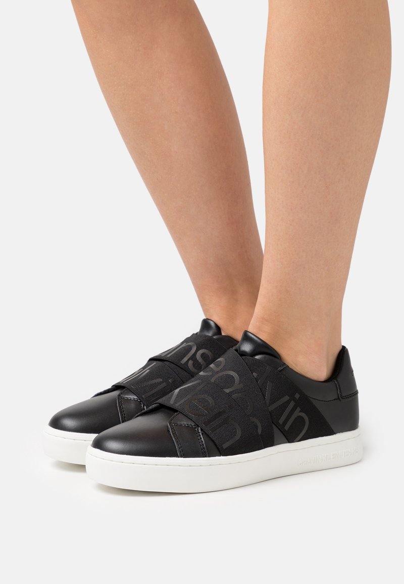 Zwarte leren sneakers met elastische banden en een logo, contrasterend met een witte rubberen zool. Luxe textuur en minimalistisch ontwerp.
