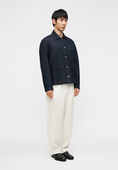 Τζιν μπουφάν σε χρώμα ίντιγκο με γιακά, κούμπωμα με κουμπιά και δύο μπροστινές τσέπες, φορεμένο πάνω από λευκό λινό παντελόνι και μαύρα παπούτσια.
