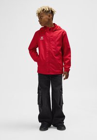 Red lightweight jacket with hood and zipper, features logo on the chest, paired with loose black cargo pants and black sneakers.