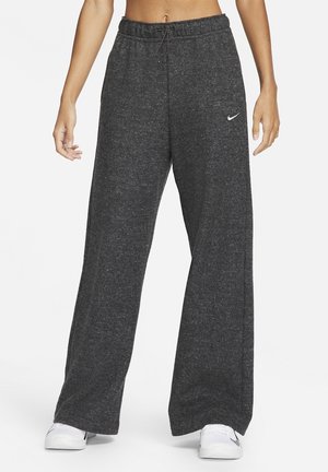 Person trägt dunkelgraue, weit geschnittene Sweathose mit elastischem Bund und weißem Nike-Logo am linken Oberschenkel, kombiniert mit weißen Sneakern.
