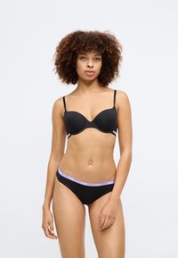 Sosten negro con copas moldeadas y tirantes delgados ajustables. Las braguitas negras a juego tienen una cinturilla púrpura con el logo. Textura suave, diseño minimalista.