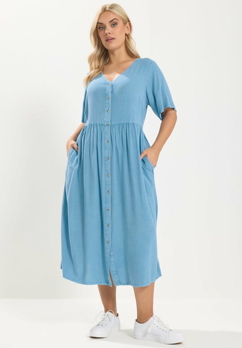 Lyshåret kvinde iført en midilang blå kjole med knapper foran, korte ærmer og hvide sneakers, hænderne i kjolens lommer.