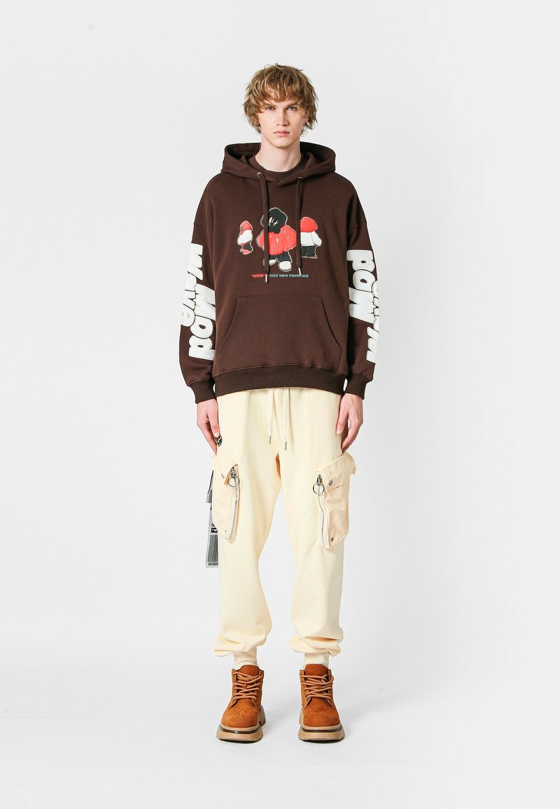 Hombre joven vestido con una sudadera gráfica marrón, pantalones cargo color crema con bolsillos grandes y botas marrón con cordones, de pie ante un fondo blanco liso.