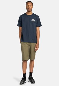 Dunkelblaues Baumwoll-T-Shirt mit weißem Logo, kombiniert mit olivgrünen Shorts. Schwarze Sportschuhe mit Mesh-Details und schwarzen Socken.