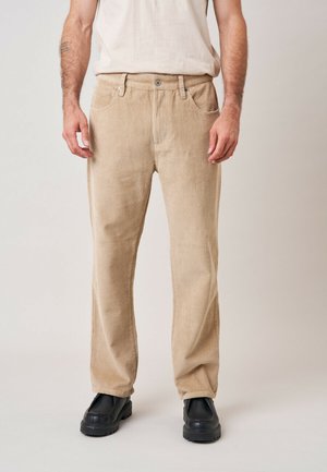 Man draagt beige ribfluwelen broek met voorzakken en zwarte schoenen tegen een effen lichte achtergrond.
