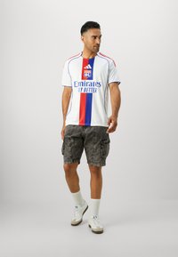 Maillot de sport blanc avec des rayures verticales bleues et rouges, arborant le logo "Emirates". Associé à un short cargo noir et des baskets blanches.