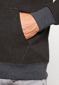 Felpa grigio scuro con tasca a marsupio e polsini a costine. Il tessuto ha una consistenza morbida con un cinturino in tonalità grigio più chiaro.