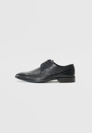 Chaussure noire en cuir avec un bout pointu, présentant un design texturé et des accents de panneaux lisses. Comprend cinq œillets pour les lacets.