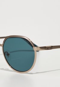 Occhiali da sole con lenti turchesi, montatura in metallo dorato e bracci con texture che presentano il logo "Guess". Forma rotonda, design elegante, hardware minimale.