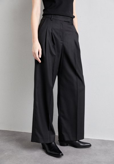 Pantaloni neri plissettati con una vestibilità sartoriale, dotati di tasche laterali e una texture liscia, abbinati a stivali neri con tacco alla caviglia.