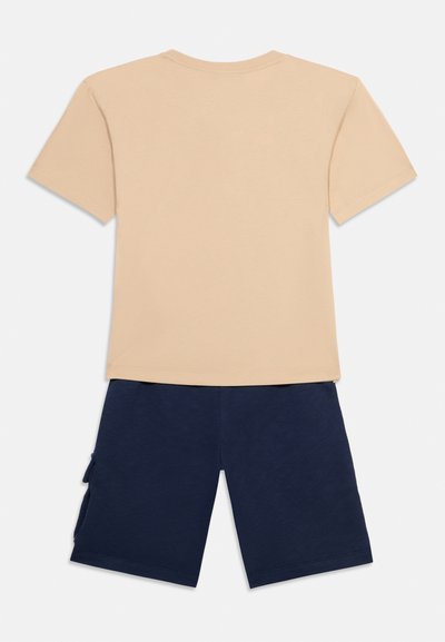 Πίσω όψη μπεζ κοντομάνικου πουκαμίσου συνδυασμένου με μπλε σκούρες βερμούδες μέχρι το γόνατο με πλαϊνές τσέπες.