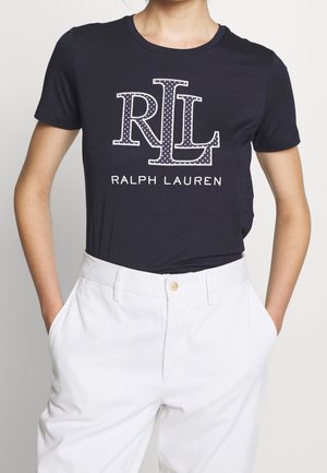 Person trägt ein dunkelblaues Ralph Lauren T-Shirt mit RL-Logo und weiße hoch taillierte Hose, Hände in den Taschen, vor neutralem Hintergrund.
