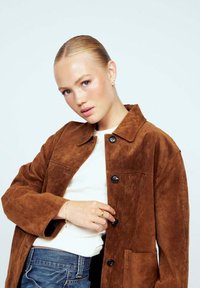 Brun mockajacka med krage, framknappar och ficka. Bärs över en vit topp och blå jeans, med en jämn textur.