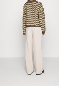 Žena v béžových širokých kalhotách a hnědo-krémovém vzorovaném pleteném svetru, stojící zády k fotoaparátu na jednobarevném pozadí.