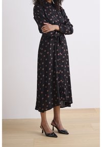 Svart klänning med vita prickar, knapp-up front, långa ärmar och ett knyt i midjan. Har en flödig silhuett och knälång fåll. Svarta högklackade skor.