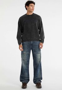 Donkergrijze trui met geribbelde boorden en marineblauwe tekst. Loszittende blauwe cargobroek met meerdere zakken en vervaagde plekken. Bruine schoenen.