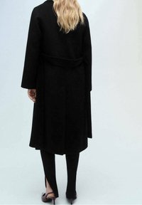 Manteau en laine noire avec une taille ceinturée, des manches longues et une coupe droite. Associé à un pantalon noir avec des fentes à l'ourlet.