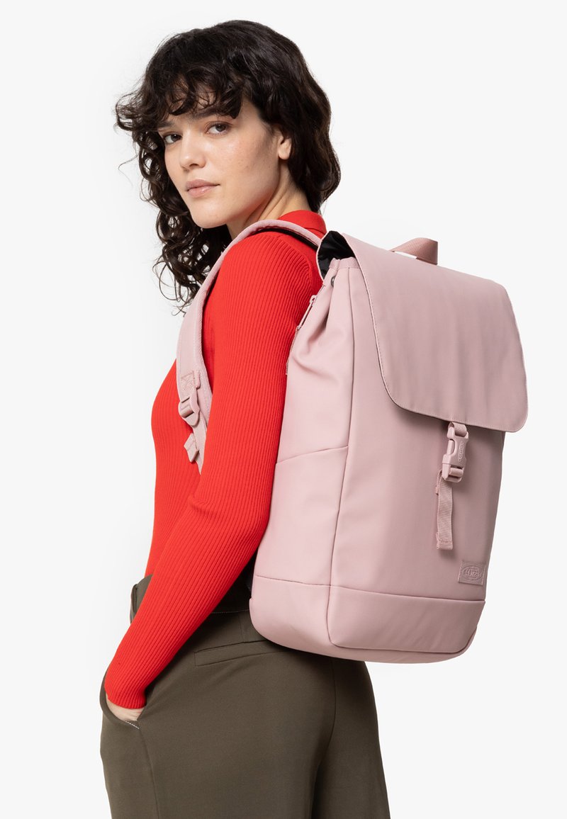 Pink backpack with a fold-over flap, adjustable straps, and minimalistic design. Material appears soft with a smooth texture.