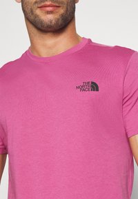 Růžové bavlněné tričko s krátkými rukávy a kulatým výstřihem. Obsahuje černé logo v oblasti horního levého hrudníku. Měkká textura.