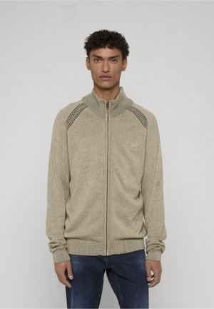 Man met kort krullend haar, gekleed in een beige gebreide trui met ritssluiting en donkere streepdetails op de schouders, en een blauwe spijkerbroek, kijkt vooruit.