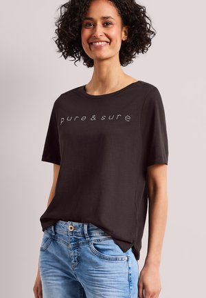 Femme aux cheveux bouclés portant un t-shirt noir avec le texte « pure & sure » et un jean bleu clair, souriante sur un fond uni.