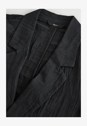 Chaqueta negra texturizada con un acabado suave, que presenta un cuello con muesca y un diseño de abertura frontal. Etiqueta de la marca "NEXT" visible en el interior.