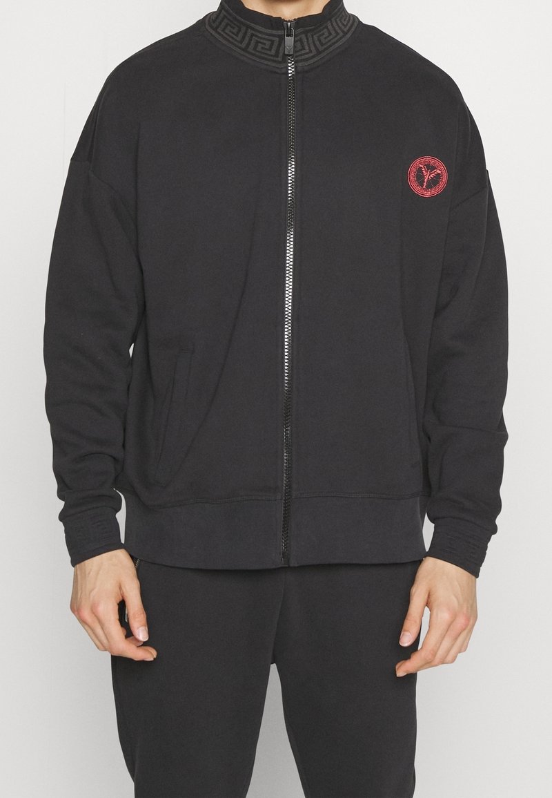 Schwarze Zip-Up Sweatjacke mit einem gemusterten Kragen, Fronttaschen und einem roten, runden Logo auf der linken Brust. Aus einem weichen Material gefertigt.