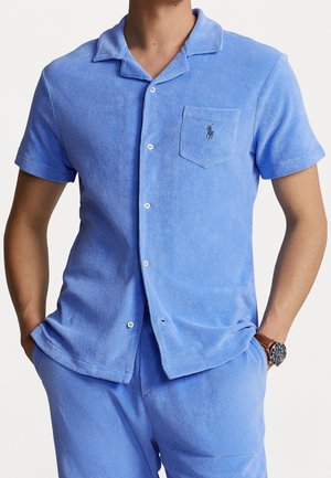 Man wearing a light blue short-sleeve terry cloth button-up shirt and matching shorts with hands in pockets and a wristwatch.