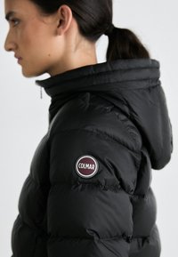 Zwarte donsjack met een hoge kraag, gestructureerde quilt en een ronde Colmar-logo patch op de linker mouw.