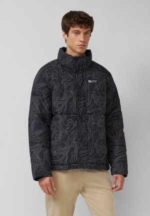 Junger Mann trägt eine schwarze Steppjacke mit abstrakten weißen Konturlinien und beige Hose, steht vor einem schlichten hellgrauen Hintergrund.