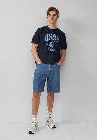 Marineblauwe T-shirt met grafische print, gecombineerd met denim shorts en lichte sneakers. Geaccentueerd met gestreepte sokken, een eenvoudig ontwerp en een ontspannen pasvorm.