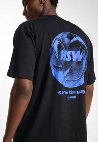 Zwarte katoenen T-shirt met een blauwe grafiek van een voetbal en de tekst "HSW" en "Serve the game" op de achterkant, met korte mouwen.