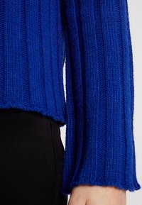 Blå stickad tröja med ribbat mönster, långa ärmar och en kortad nederkant. Tyget ser mjukt och något lurvigt ut. Svarta byxor syns.