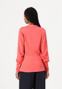 Blusa corallo a maniche lunghe, texture liscia, vestibilità morbida, con scollatura rotonda e maniche arricciate, abbinata a pantaloni scuri.