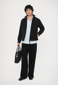 Čierna bundá na zips cez svetlomodrú košeľu, skombinovaná s voľnými čiernymi nohavicami a čiernymi topánkami. Drží čiernu tašku so strieborným kovovým hardvérom.