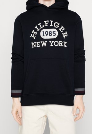 Sweat à capuche noir avec poche frontale et texte brodé en blanc "HILFIGER 1985 NEW YORK", porté avec un pantalon de couleur claire.