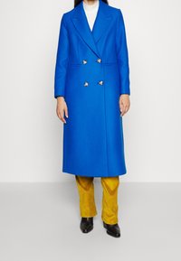 Cappotto doppiopetto blu in lana con bottoni argentati, dotato di colletto a revers. Abbinato a pantaloni in velluto a coste gialli e stivali neri alla caviglia.