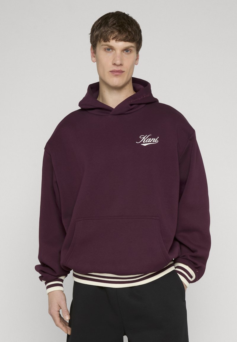 Junger Mann, der einen kastanienbraunen Hoodie mit weißem "Kani"-Logo, gestreiften Bündchen und Saum trägt, steht vor einem schlichten hellgrauen Hintergrund.