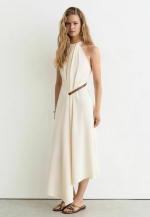 Junge Frau mit langen blonden Haaren, die ein cremefarbenes ärmelloses Neckholderkleid mit braunem Gürtel und flachen Sandalen trägt und vor einer schlichten Wand steht.