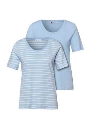 Unge kvinde iført en lyseblå og hvidstribet T-shirt og blå jeans, stående mod en ensfarvet lys baggrund.