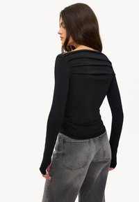 Zwarte langiemouwige top met geplooide details op de rug, gecombineerd met grijze hoge taille jeans, weergegeven vanuit een achteraanzicht.