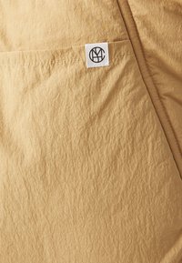 Tissu froissé beige avec une poche comportant une petite étiquette blanche rectangulaire affichant un logo circulaire noir.