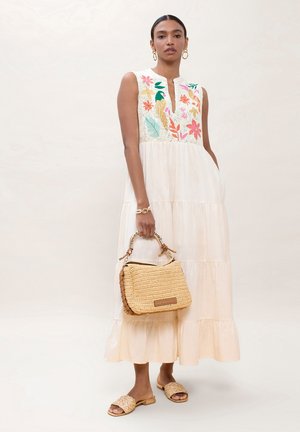 Mujer con vestido sin mangas color crema con bordado floral colorido, llevando un bolso tejido y sandalias deslizadas a juego también tejidas.