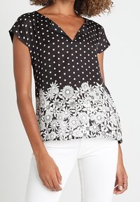Černé tričko s krátkým rukávem, zdobené bílými puntíky a květinovým vzorem na lemu. Hladká látka s výstřihem do V a krátkými rukávy.