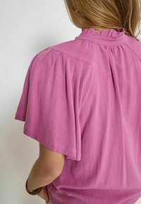 Blusa rosa a maniche corte con scollo a balze, tessuto morbido e dettagli plissettati sulle spalle. Design semplice e dal taglio rilassato.