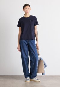 Marinblå bomullströja med korta ärmar och vitt broderat logotyp, kombinerad med lösa blå denimjeans och grå sneakers, hållandes en beige väska.