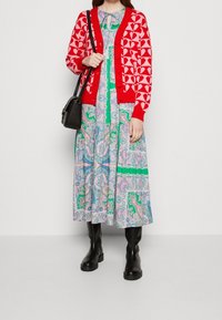 Červený cardigan s vzorem srdcí, vrstvený přes světle modré šaty s paisley potiskem, které mají zelené a růžové akcenty, doplněný černými kozačkami po kolena.