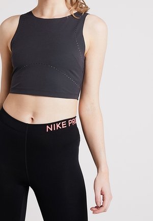 Vrouw draagt een zwart, mouwloos crop top en zwarte Nike Pro leggings, staand met één arm ontspannen langs haar lichaam.
