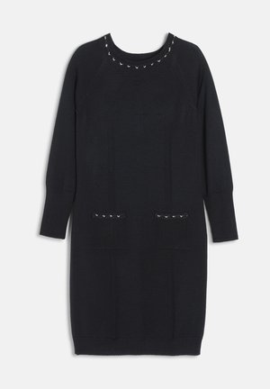 Femme portant une robe en maille noire à manches longues avec des détails à clous, des talons noirs et un sac à épaule noir avec une fermeture dorée, debout devant un fond blanc.